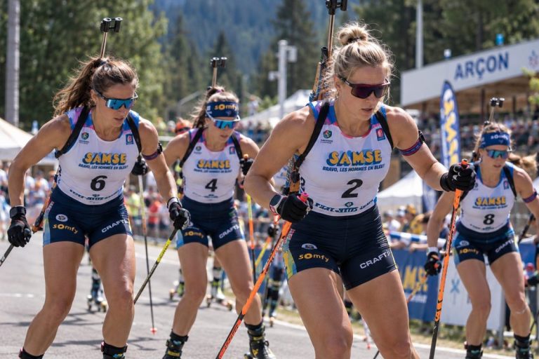 Biathlètes féminines en pleine course de ski-roues lors du Summer Tour d’Arçon 2026, avec carabines sur le dos, tenues de compétition et spectateurs au bord du parcours.