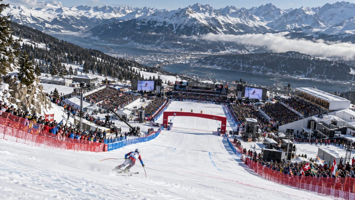 Vue plongeante de l’aire d’arrivée d’une piste de descente lors d’une grande compétition de ski alpin, avec un skieur en approche, des tribunes remplies de spectateurs et les montagnes enneigées en arrière-plan.