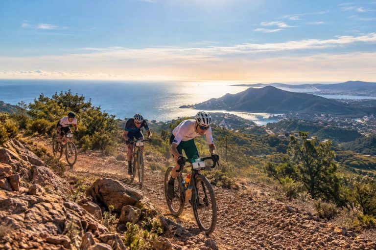 Trois coureurs en gravel gravissent une piste rocailleuse sur les hauteurs de la Côte d’Azur, lors d'une course de vélo en terrain montagneux.