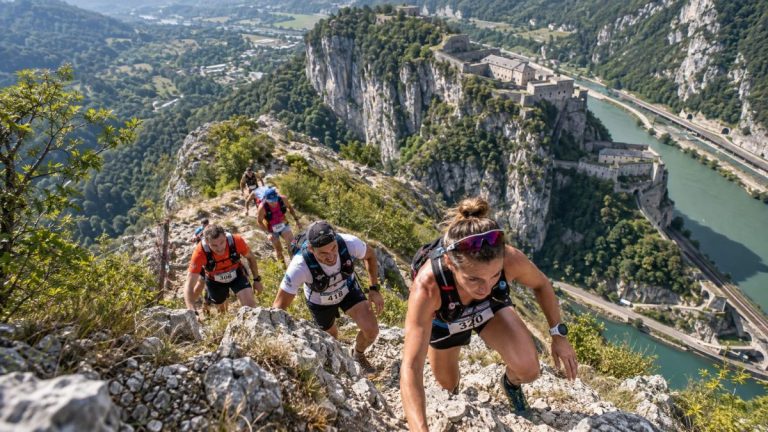 Traileurs et traileuses vus de haut en plein effort sur une arête rocheuse au Défi du Fort l’Écluse 2026, avec le fort perché sur la falaise et le Rhône en contrebas.
