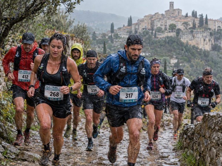 Coureurs et coureuses participant au Trail de Tourrettes-sur-Loup 2026 sous une pluie battante, avec des tenues de trail trempées, des dossards et le village perché en arrière-plan